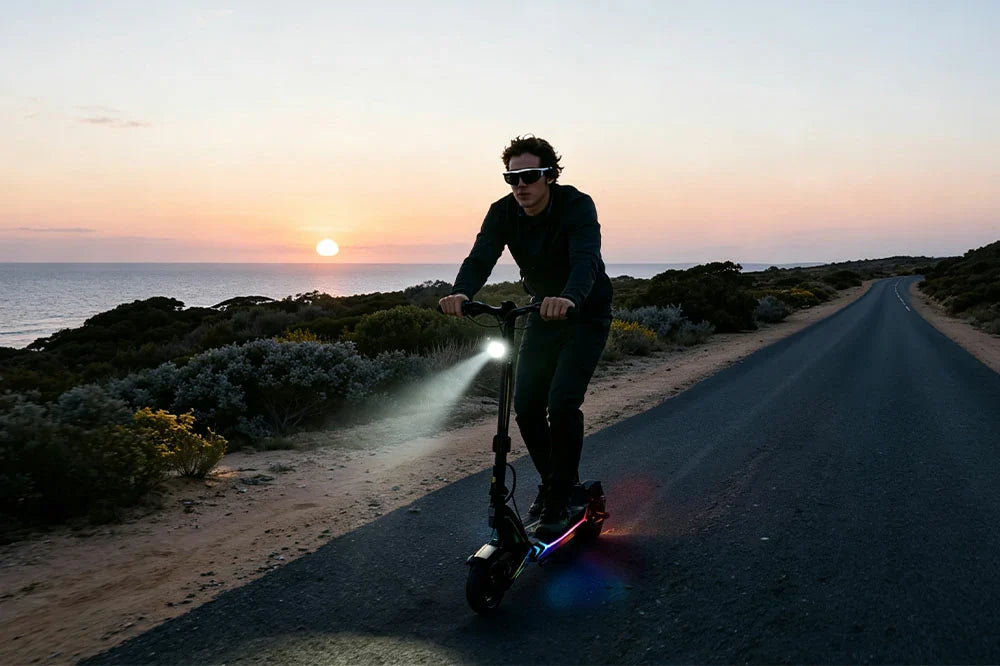 Male rider riding RGB illuminated electric scooter on coastal asphalt road at sunset, ocean and dusk mountain backdrop, bright front headlight