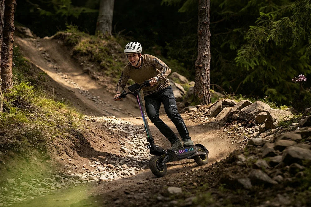 Male rider with helmet riding RGB lighted off‑road electric scooter on rough mountain dirt trail, extreme outdoor sport e‑scooter